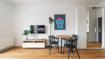 Ein modernes Wohnzimmer mit Fernseher, Couchtisch und vier Stühlen. Auf einem Beistelltisch steht eine Pflanze, an der Wand hängt ein Gemälde, und eine Tür führt in einen anderen Raum.