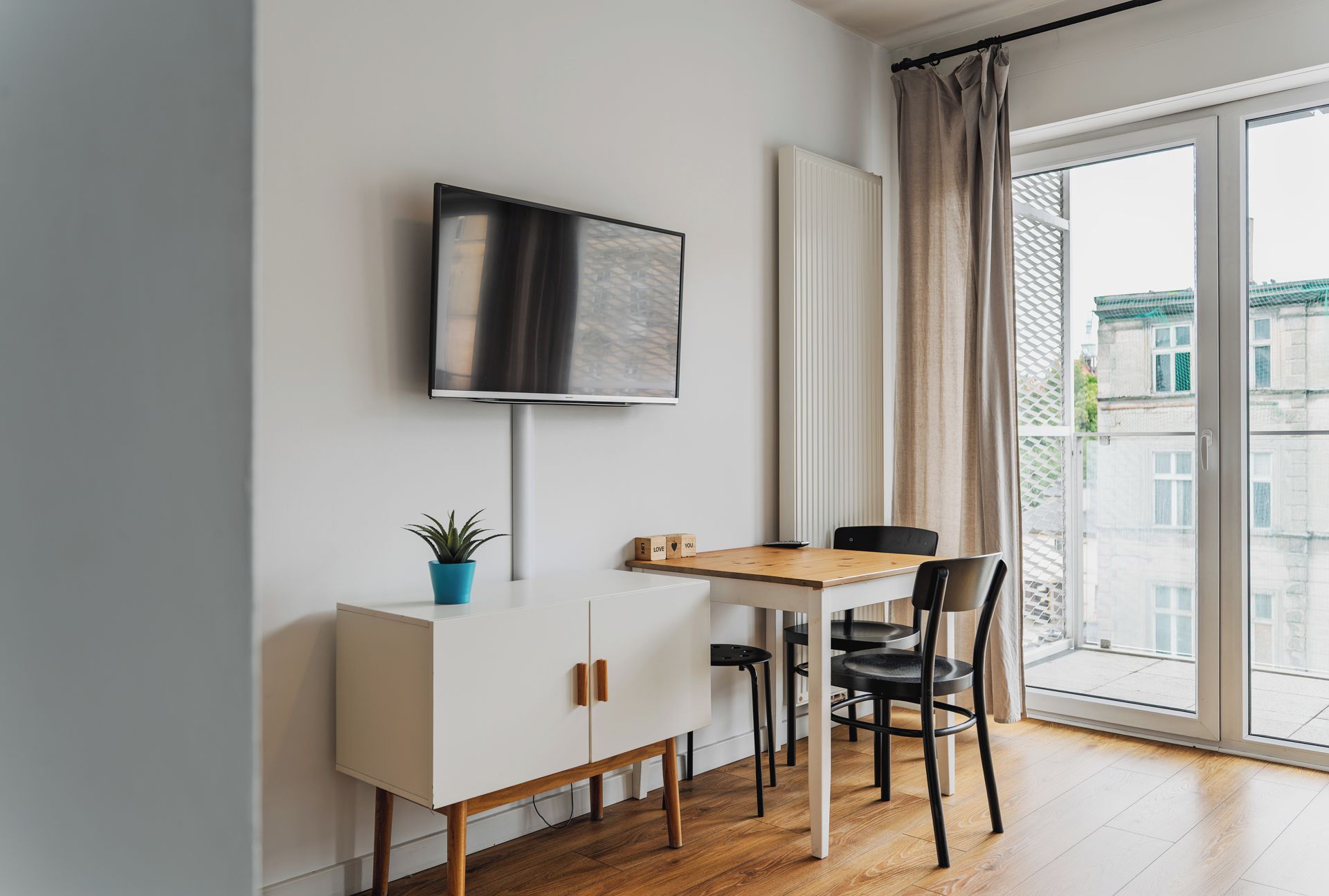 A modern living space with a flat-screen TV mounted on the wall, a wooden dining table with chairs, a white cabinet, and a potted plant.
