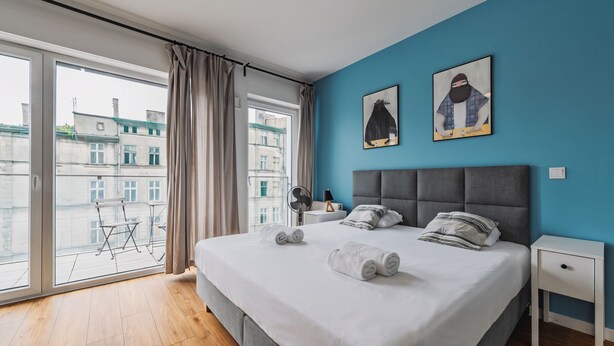 Ein Hotelzimmer mit einem großen Bett, zwei Wandbildern, Blick auf Gebäude durch das Fenster und einem Balkon mit Tisch und Stühlen.