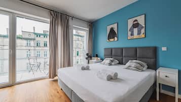 Habitación de hotel con una cama grande, dos obras de arte en la pared, vista a edificios por la ventana y un balcón con mesa y sillas.