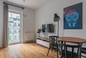 Una sala de estar moderna con suelo de madera, una mesa redonda, una silla negra, un televisor sobre un mueble bajo, un radiador y una ventana con cortinas.