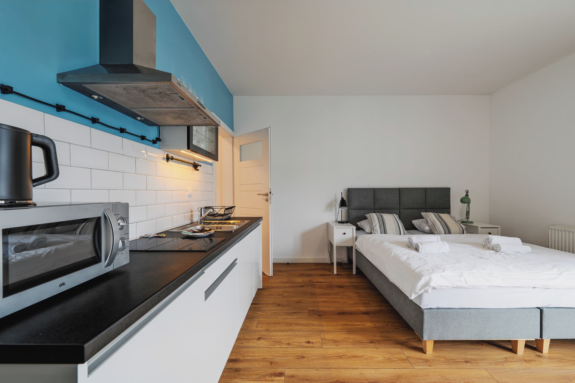A compact hotel room with a kitchenette, a bed with grey bedding, and a wooden floor.