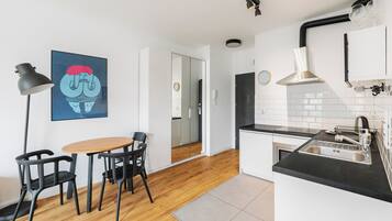 A modern kitchen with a black countertop, stainless steel sink, and a range hood. There is a dining area with a table and chairs, and a black floor lamp.