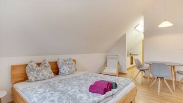 A bedroom with a bed, a chair, a small table, and a kitchenette in the background.