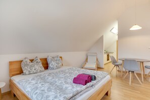 A bedroom with a bed, a chair, a small table, and a kitchenette in the background.
