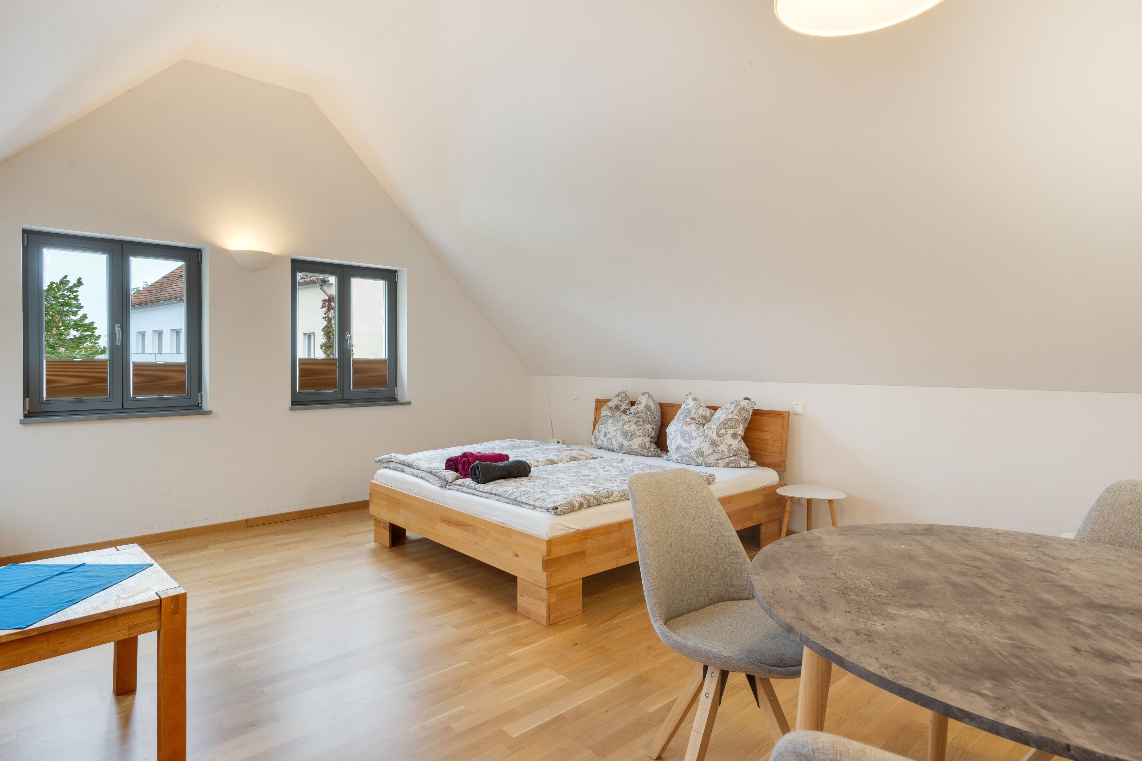 Un dormitorio con techo inclinado, cama de madera, una mesita y una silla.