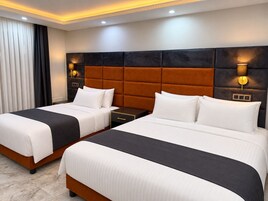 Habitación de hotel con dos camas, cada una con ropa de cama blanca y cabeceros oscuros. Se observan mesitas de noche con lámparas, y la habitación presenta un diseño moderno con suelo de mármol.