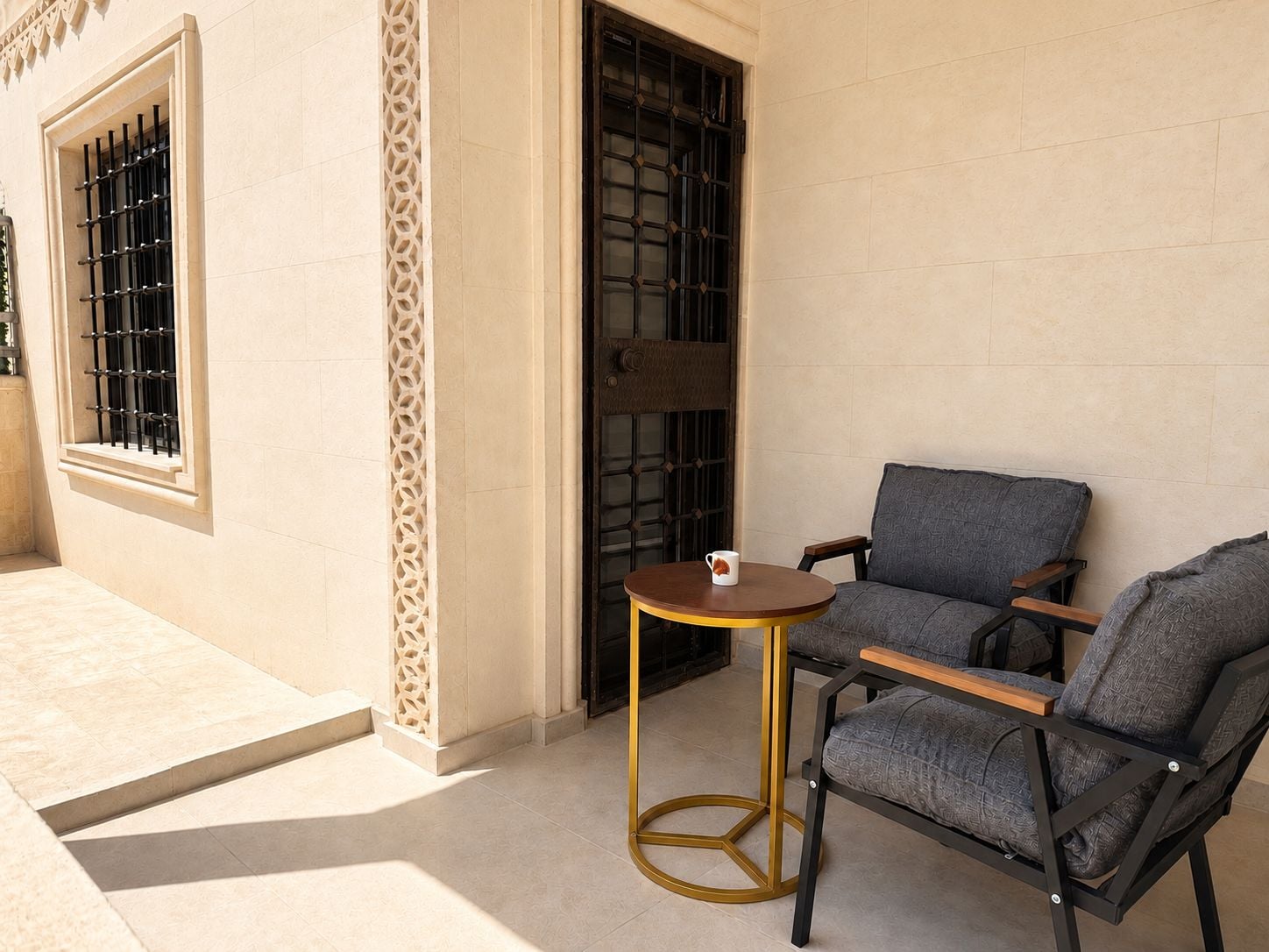 An outdoor seating area with a round table and two chairs, a window with metal bars, and a decorative wall panel.