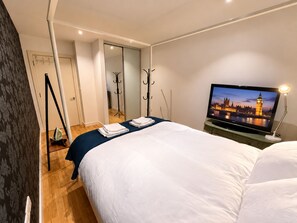 Et hotelværelse med en seng, et fjernsyn der viser et bybillede, et skrivebord med en lampe og et knagered.