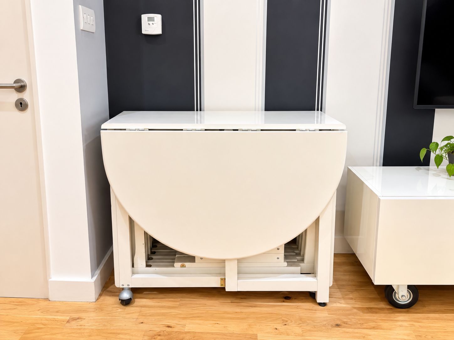 A white, semi-circular, wheeled table with a flat top, positioned in a room with wooden flooring, a door, and a wall-mounted thermostat.