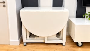 Una mesa blanca, semicircular y con ruedas, con una superficie plana, ubicada en una habitación con suelo de madera, una puerta y un termostato fijado a la pared.