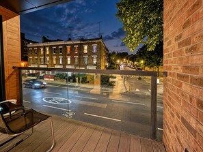 Un balcón con vista a una calle por la noche, que incluye un edificio de ladrillo y coches estacionados.