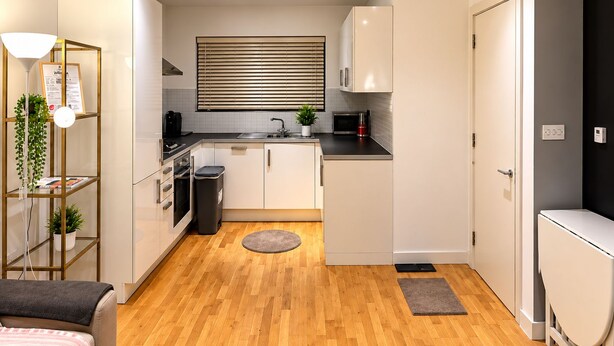 Een kleine keuken met witte kastjes, een zwart aanrecht en een raam met zonwering.