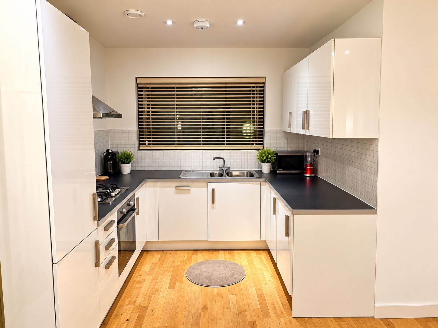 A modern kitchen with white cabinets, a black countertop, a stainless steel sink, a window with blinds, and a wooden floor.