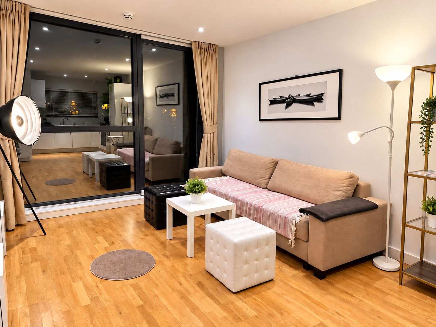 A modern living room with a sofa, ottoman, and a small table. A sliding glass door leads to a dining area with a kitchen in the background.