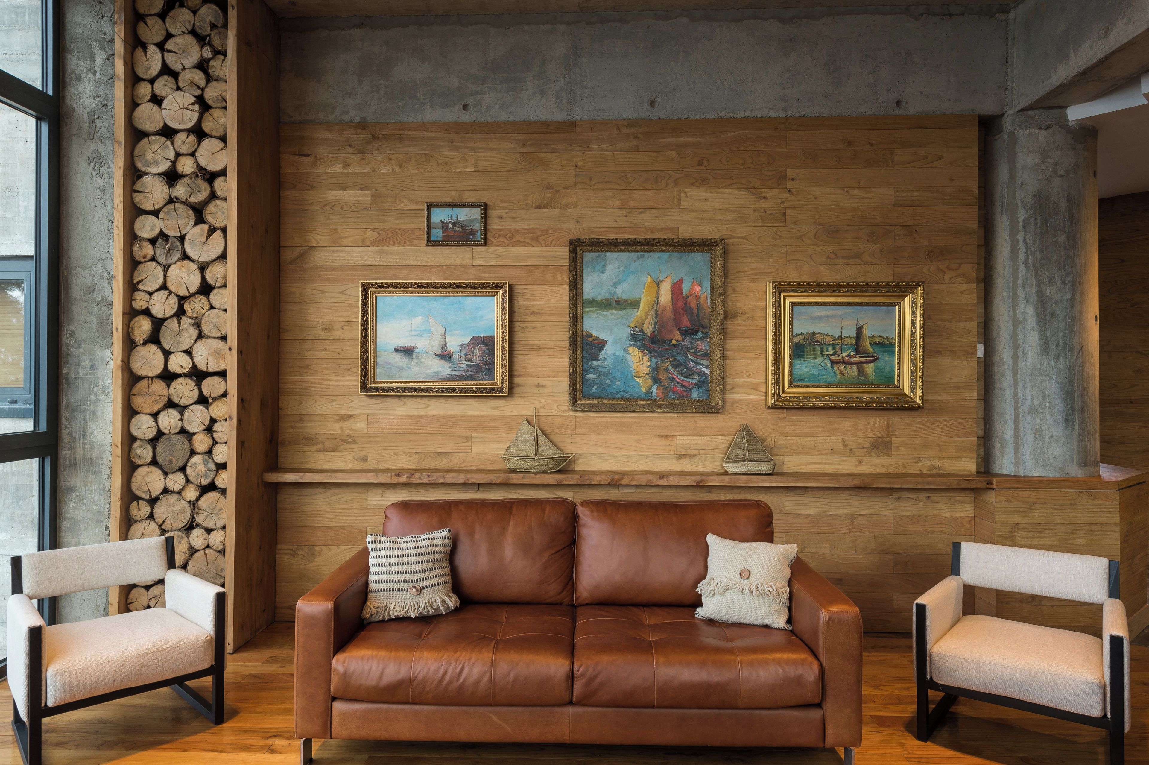 A room with a brown leather sofa, two armchairs, and three framed paintings on the wall.