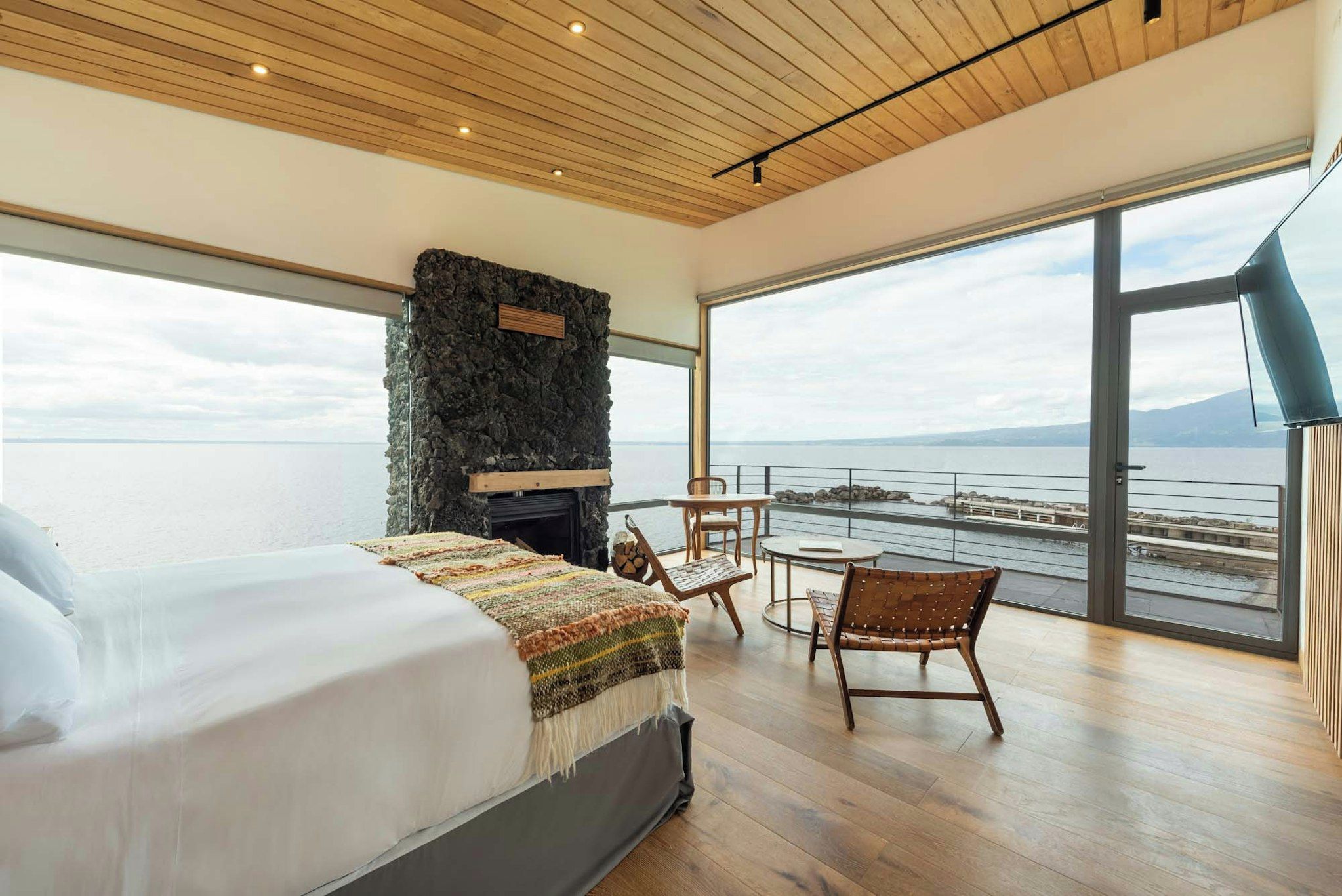 A bedroom with a large bed, a stone fireplace, a view of the sea, and a balcony with chairs.