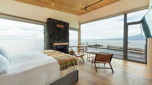 Een slaapkamer met een groot bed, een stenen openhaard, uitzicht op zee en een balkon met stoelen.