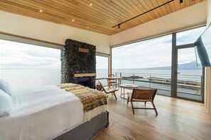 A bedroom with a large bed, a stone fireplace, a view of the sea, and a balcony with chairs. - Hotel Awa (Puerto Varas)
