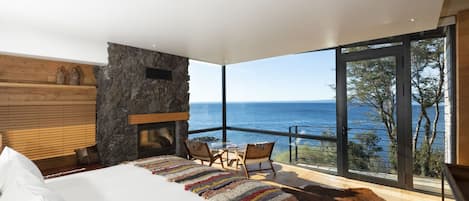 Un dormitorio con una cama grande, chimenea de piedra, vista al mar y un balcón con mesa y sillas.
