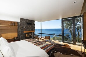 A bedroom with a large bed, a stone fireplace, a view of the sea, and a balcony with a table and chairs. - Hotel Awa (Puerto Varas)