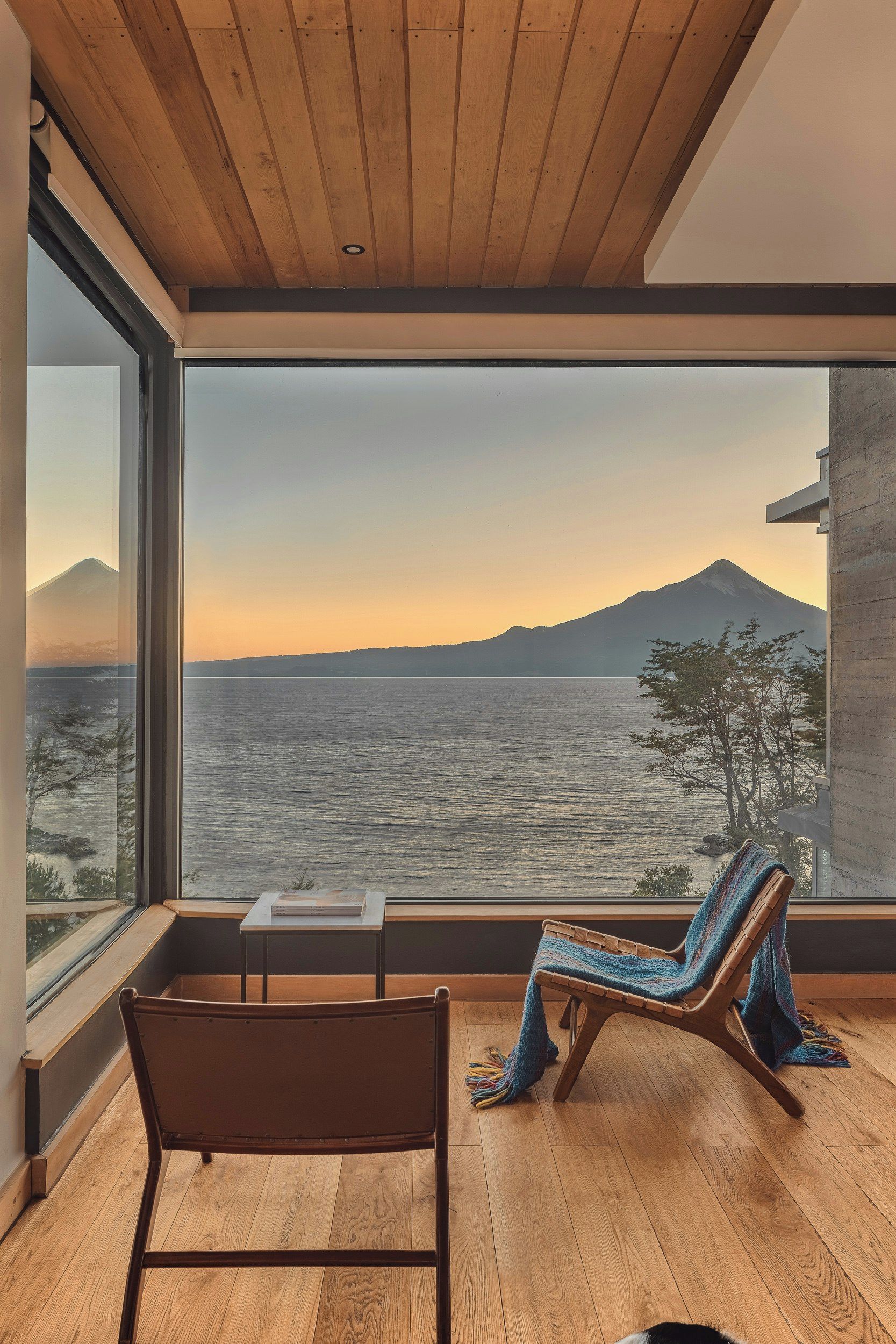 A room with a large window overlooking a body of water and a mountain. There is a wooden floor, a chair, and a small table.