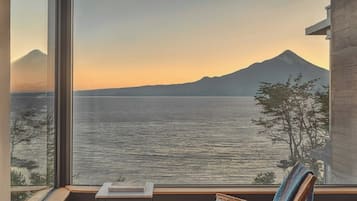Habitación con un gran ventanal con vista a un cuerpo de agua y una montaña. Tiene piso de madera, una silla y una pequeña mesa.