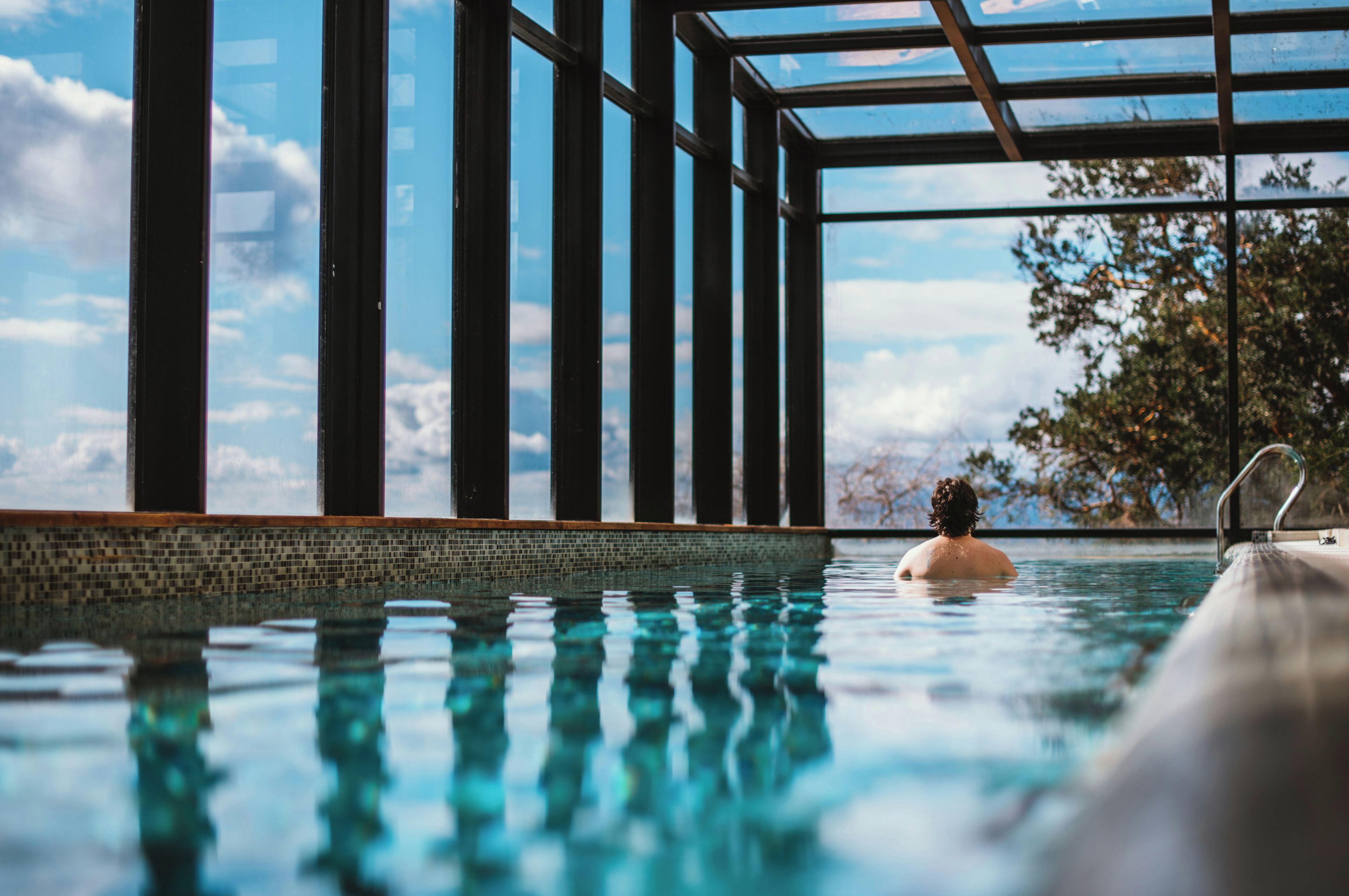 A swimming pool with clear water, surrounded by a tiled edge and a glass enclosure with metal supports.
