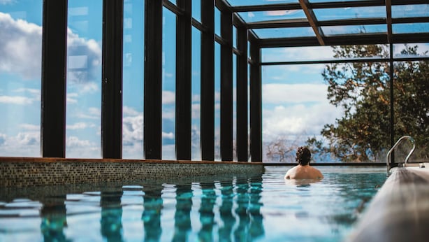 A swimming pool with clear water, surrounded by a tiled edge and a glass enclosure with metal supports. - Hotel Awa (Puerto Varas)