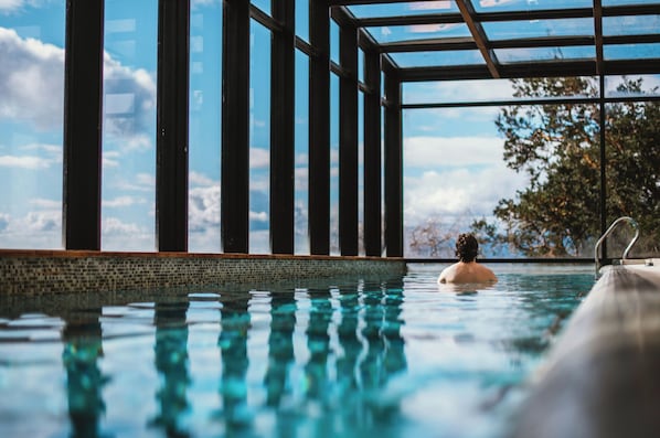 A swimming pool with clear water, surrounded by a tiled edge and a glass enclosure with metal supports. - Hotel Awa (Puerto Varas)