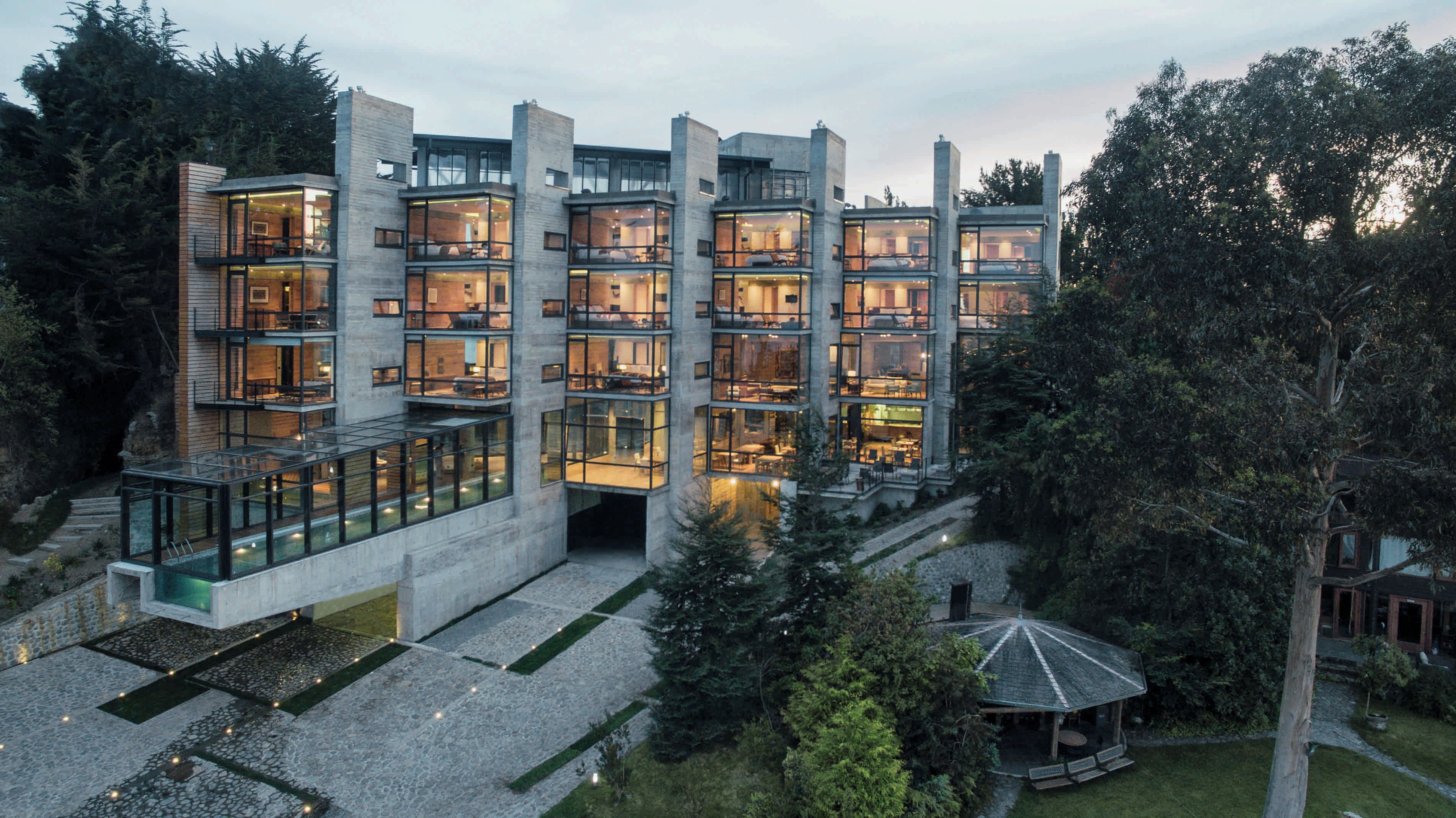 A multi-story building with large windows and a glass facade, surrounded by trees and a small gazebo.
