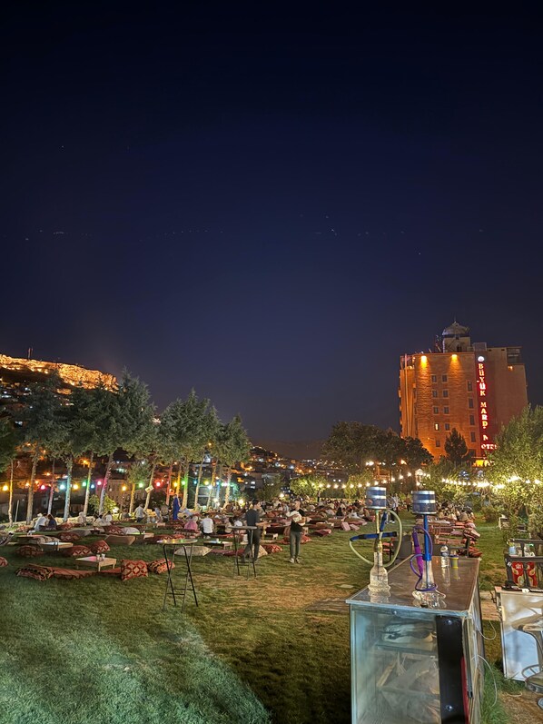 An outdoor seating area with tables and chairs, a stage, and a large building in the background. - Buyuk Mardin (Mardin)