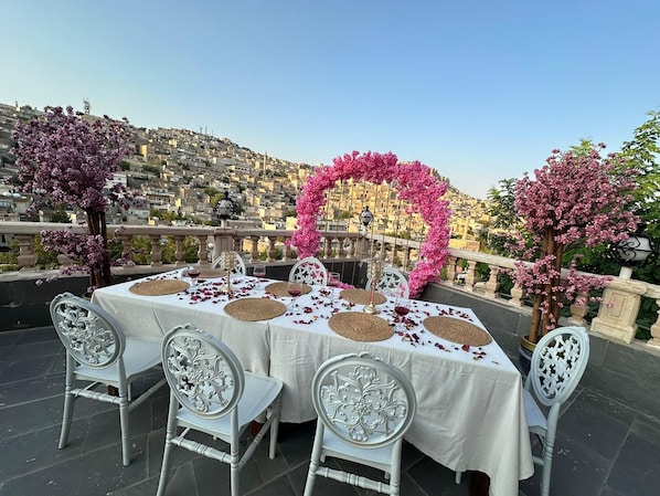 Ein Essbereich im Freien mit einem Tisch für vier Personen, umgeben von weißen Stühlen, auf einer Terrasse mit Blick auf eine dicht bebunte Stadtlandschaft.