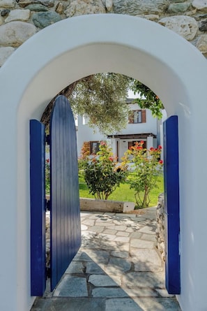 Un sendero de piedra conduce a un edificio blanco con puerta azul, rodeado de vegetación y flores.