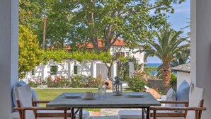 Ein Essbereich im Freien mit einem Tisch für zwei Personen, umgeben von weißen, gepolsterten Stühlen, mit Blick auf einen Garten, Bäume und im Hintergrund Gebäude.