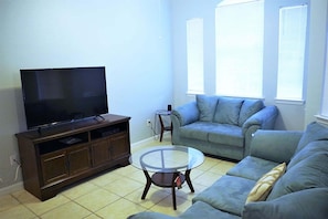 Una sala de estar con una televisión de pantalla plana, una mesa de centro de vidrio, un sofá y una mesita auxiliar.