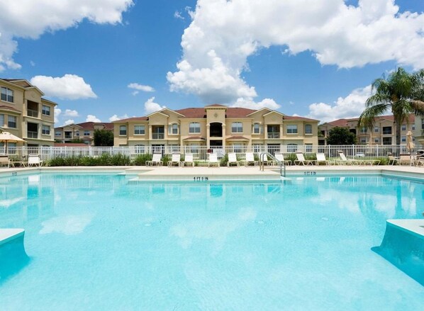 Una piscina con valla blanca, sillas de descanso y una hilera de edificios al fondo.