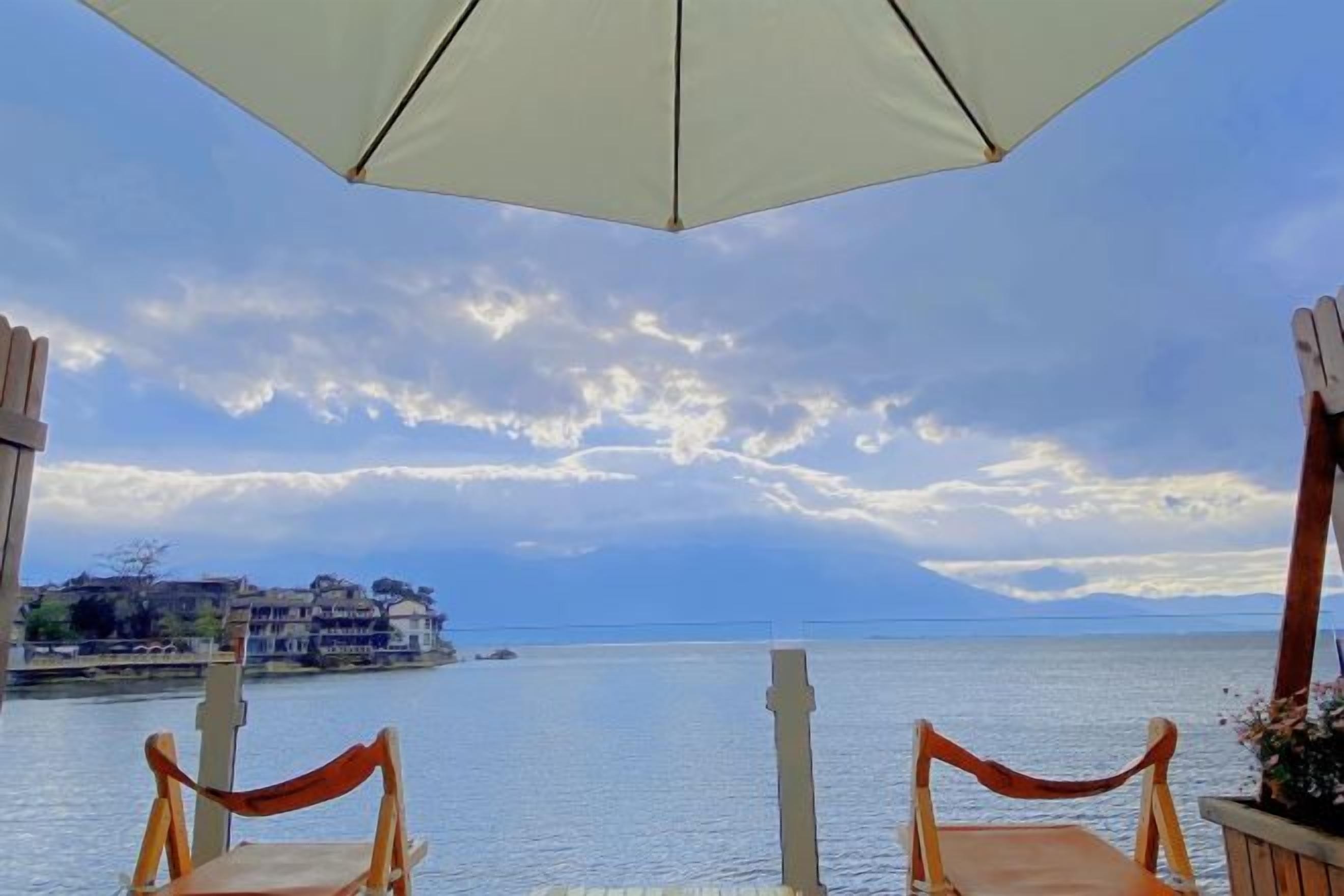 Vista al mar con cielo despejado, dos sillas de madera bajo una sombrilla blanca y un mar tranquilo.