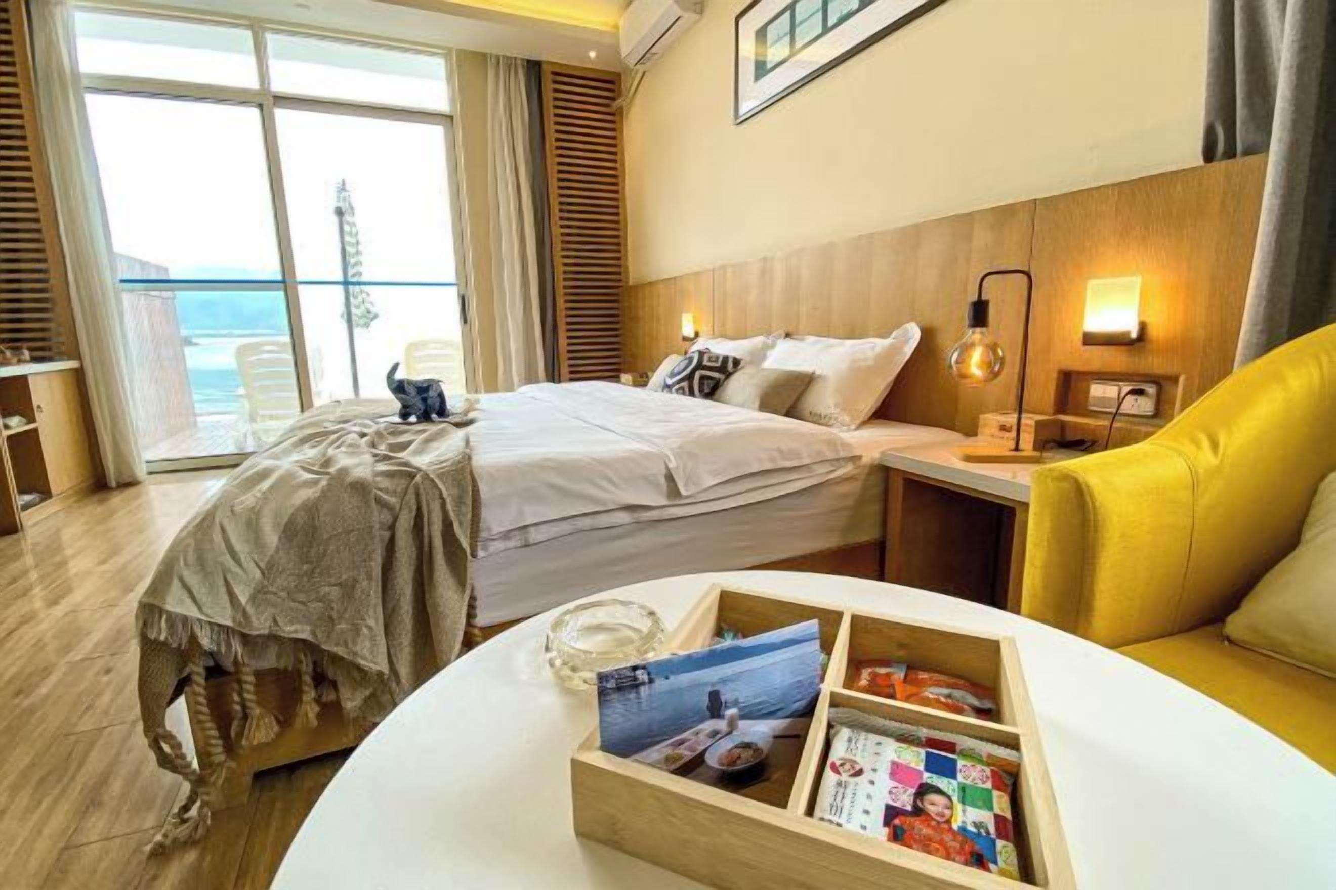 Ett hotellrum med en säng, ett sängbord, en gul fåtölj och utsikt över havet genom stora fönster.