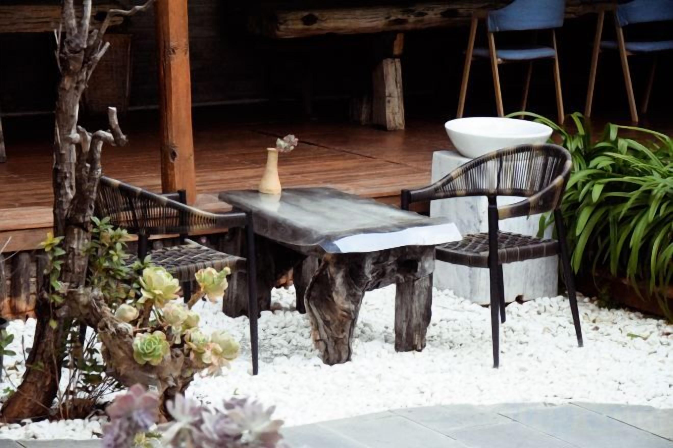 Un espacio al aire libre con una mesa y sillas de madera rústica, rodeado de piedras blancas y follaje.