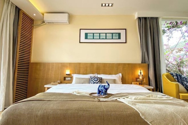 A hotel room with a large bed, a bedside table with a lamp, a framed picture on the wall, and a window with curtains.