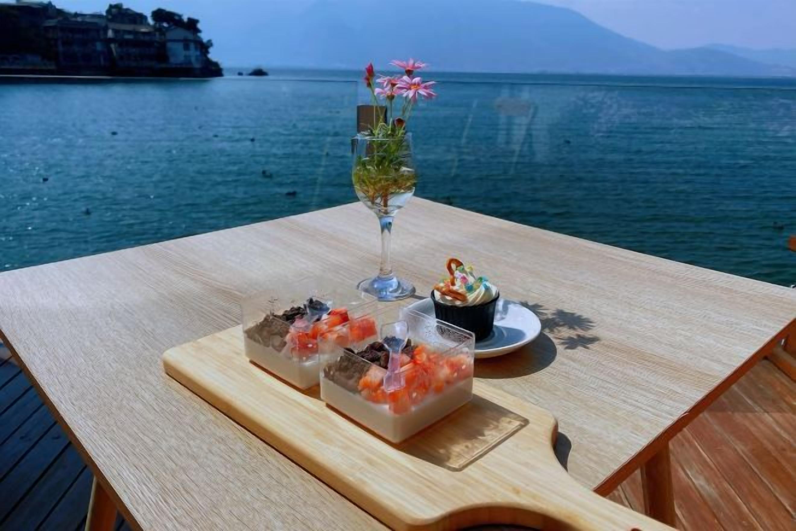 Una mesa con un vaso de bebida, un postre y una bandeja de comida, sobre un fondo de agua y montañas a lo lejos.