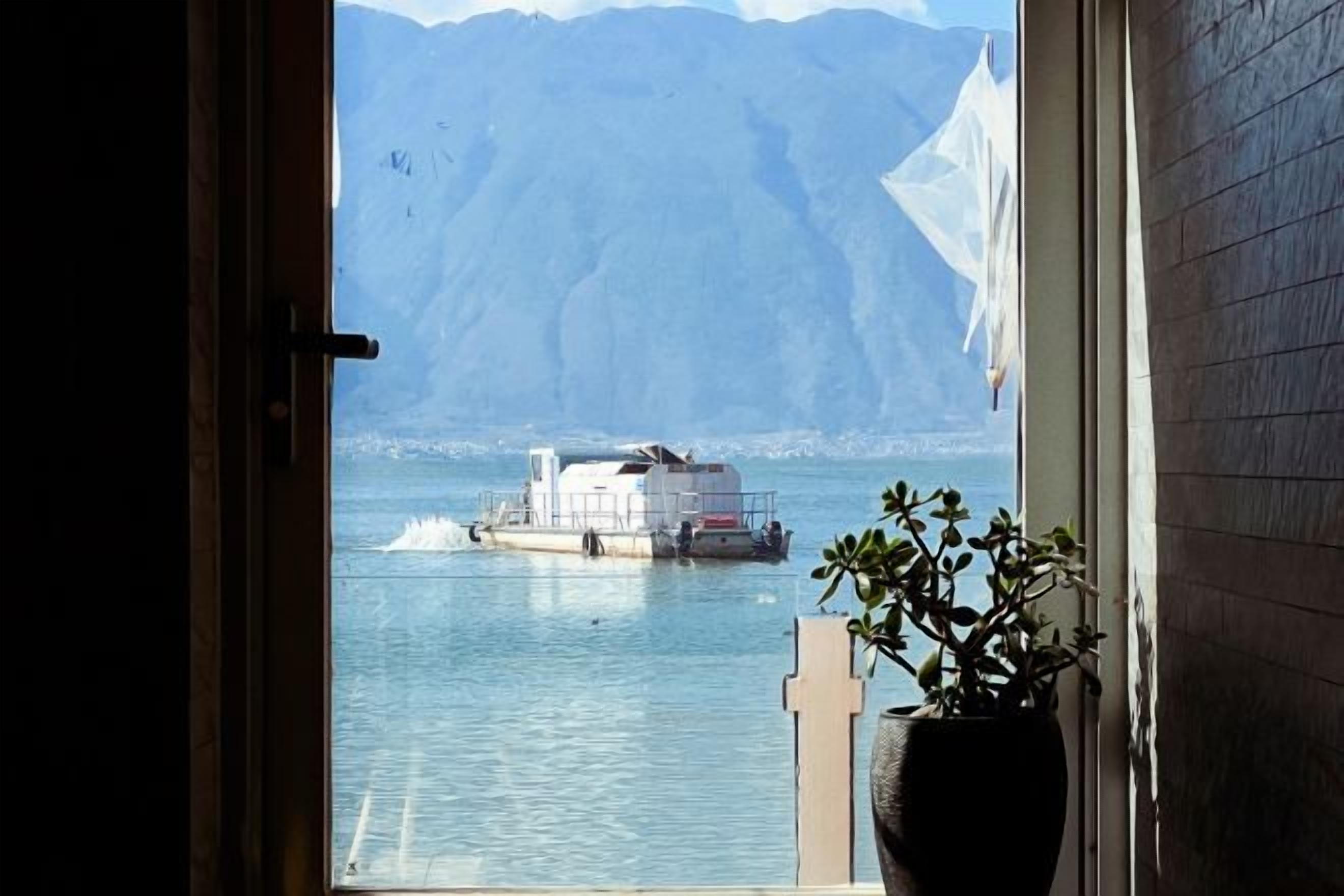Vista desde una ventana con vista a una masa de agua, un barco, un paisaje montañoso y una planta en maceta en el alféizar.