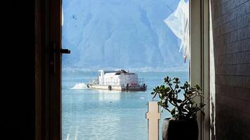 Vista desde una ventana con vista a una masa de agua, un barco, un paisaje montañoso y una planta en maceta en el alféizar.