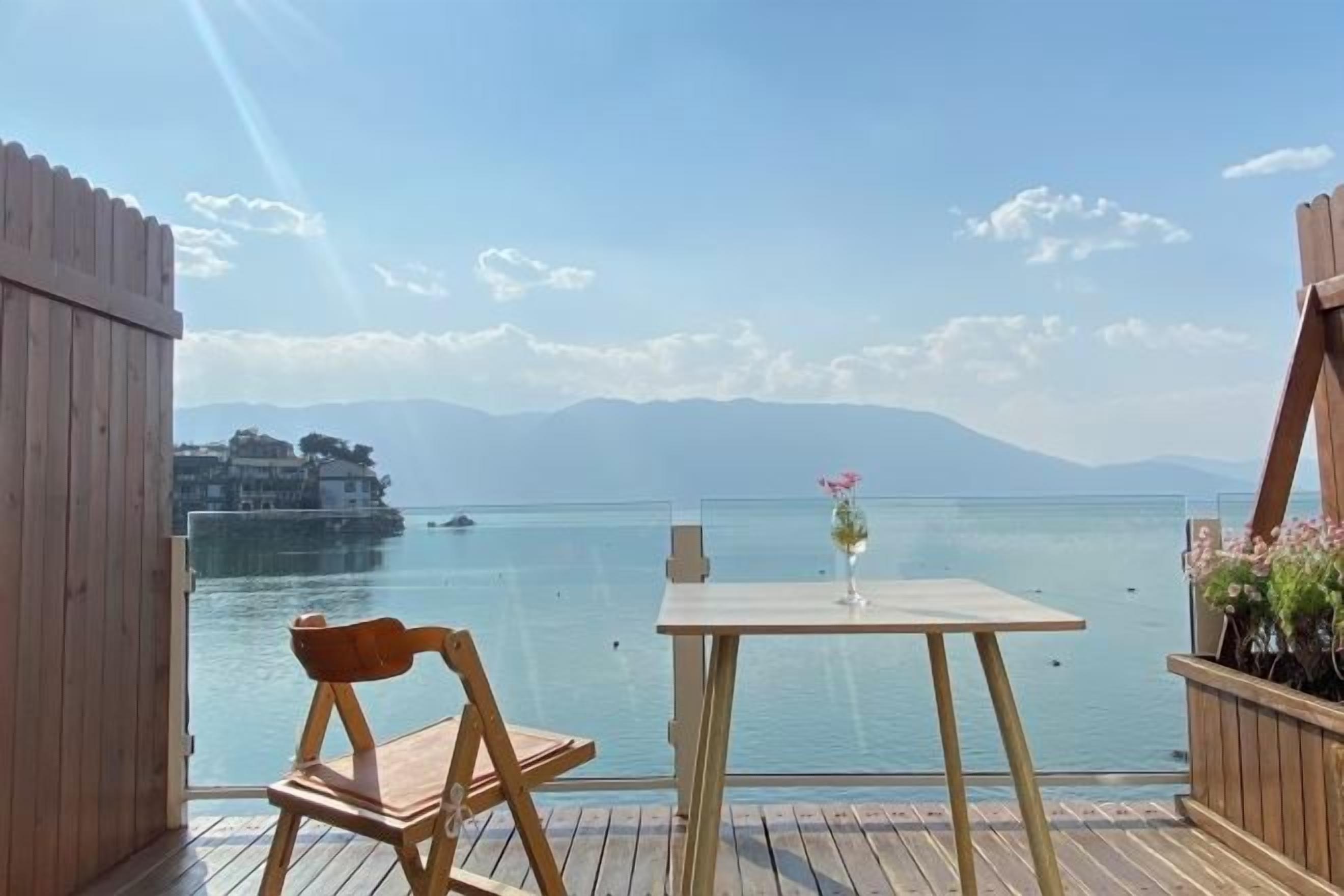 Una terraza de madera con una mesa y una silla que da a un cuerpo de agua y montañas a lo lejos.