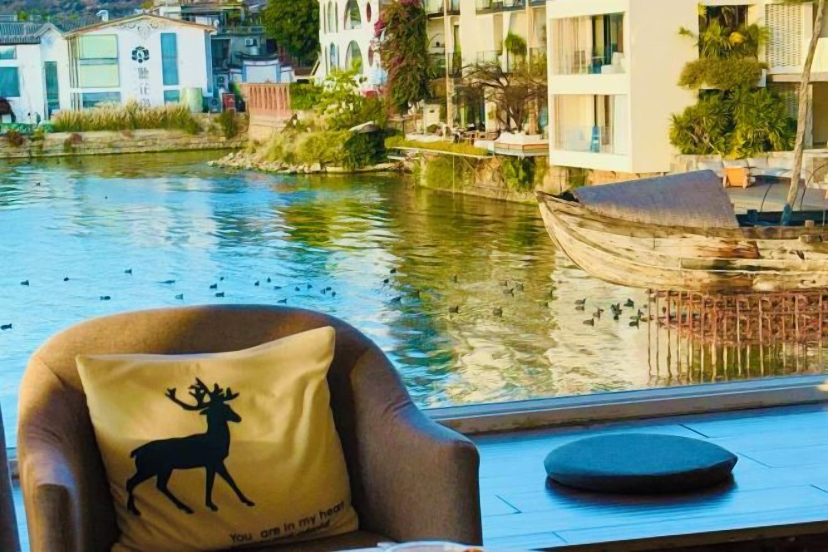 A chair with a pillow featuring a deer silhouette and text, a round table, and a view of a river with boats and buildings in the background.