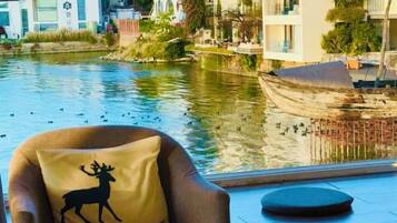 A chair with a pillow featuring a deer silhouette and text, a round table, and a view of a river with boats and buildings in the background.