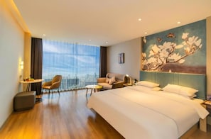 A hotel room with a large bed, a seating area with a sofa and armchairs, a small table, and a city view through large windows. - Magnotel (Jiaxing)