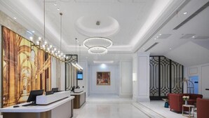 Um saguão de hotel com balcão de recepção, um lustre, uma pintura grande e uma área de estar com sofás vermelhos.