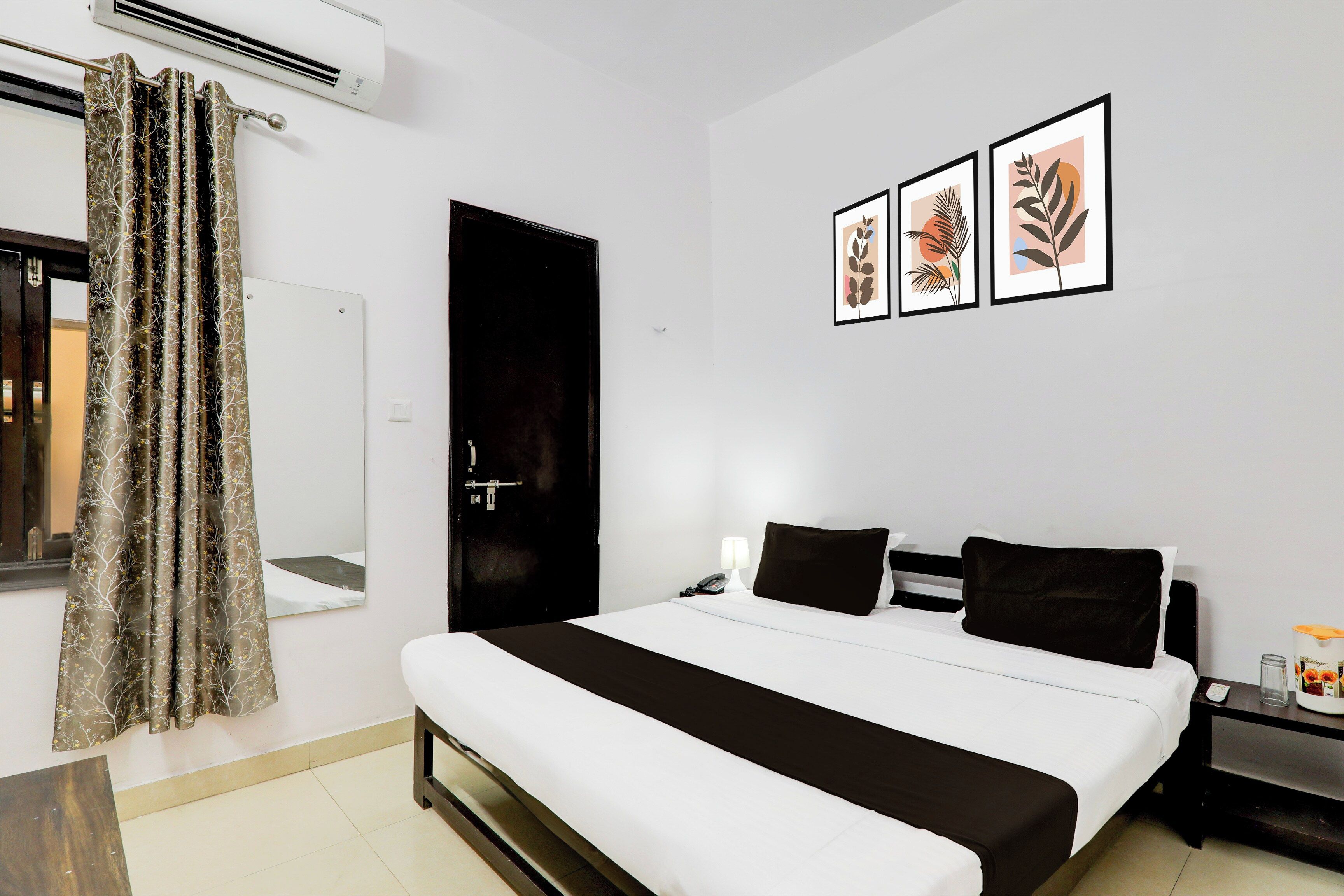 Habitación de hotel con una cama, una mesita de noche con lámpara, un teléfono y un cuadro de una planta en la pared.